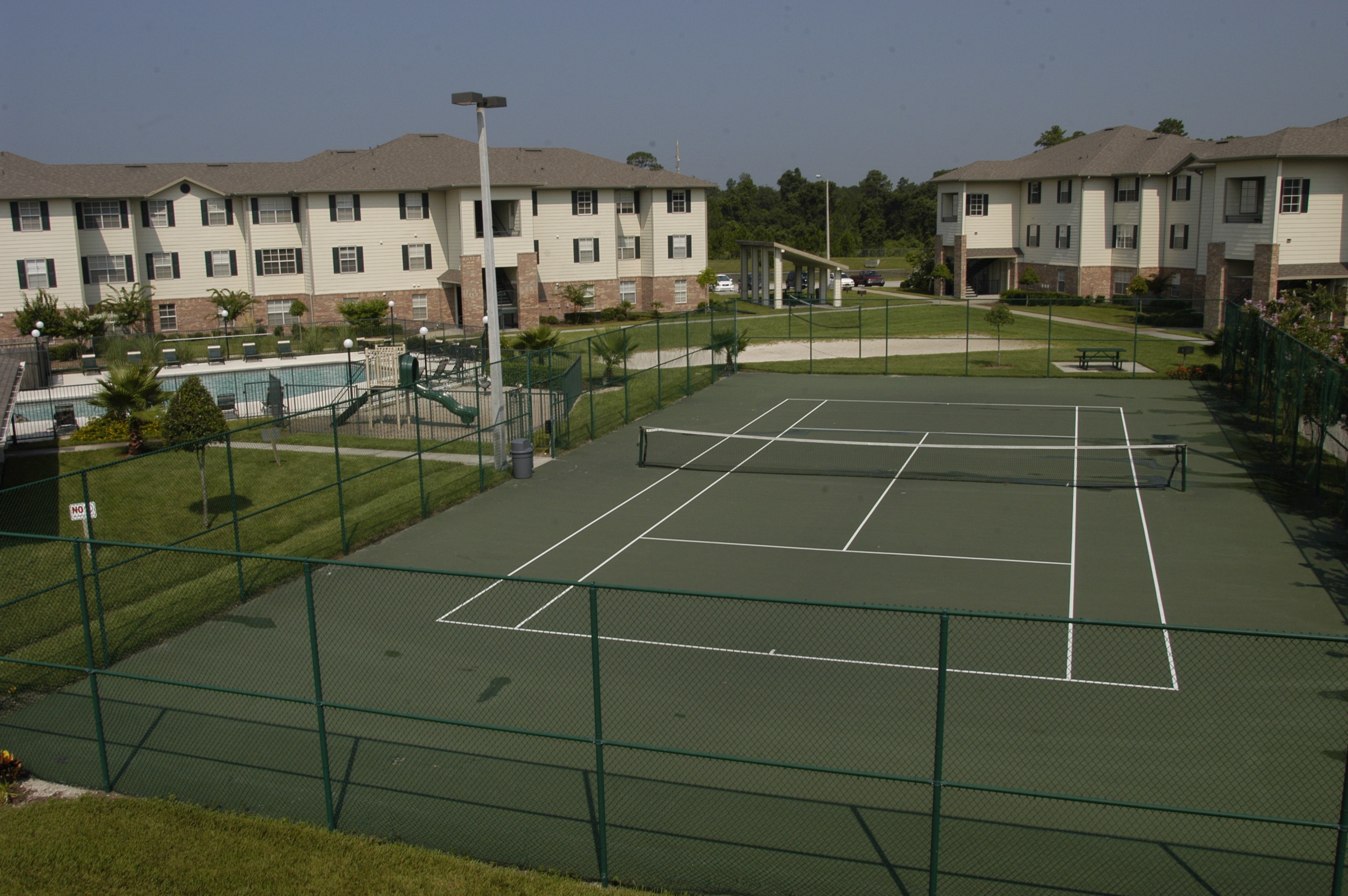 A tennis court surrounded by a green fence and apartment buildings in the background.